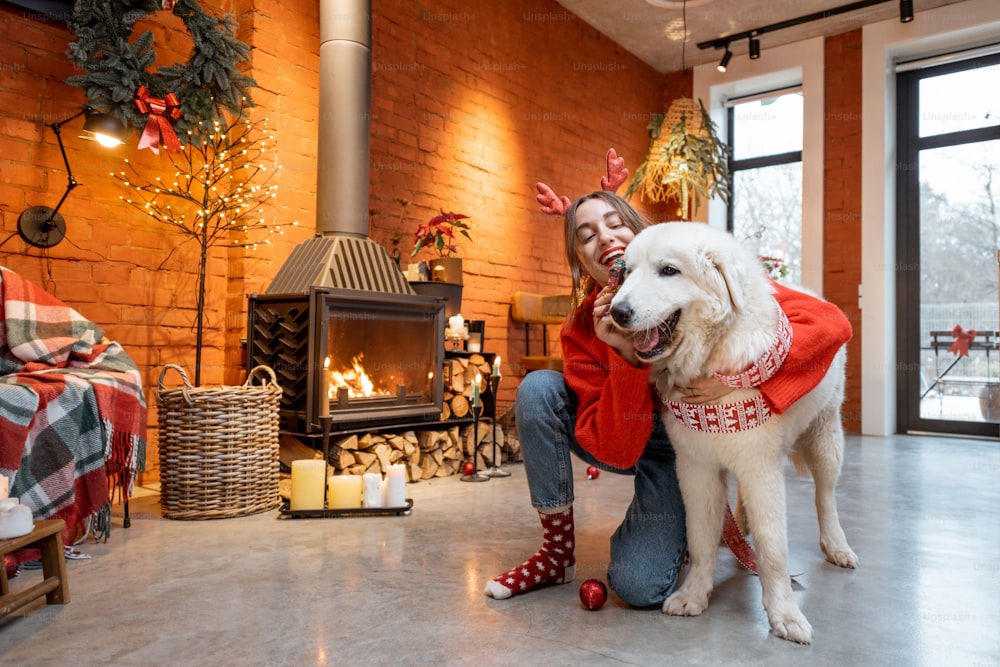 Junge Frau mit ihrem süßen weißen Hund während eines glücklichen Neujahrsurlaubs sitzt am Kamin zu Hause