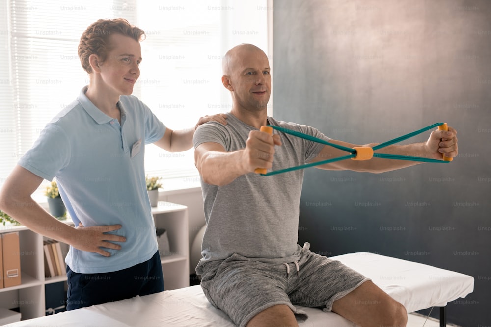Bald man stretching resistance band during exercise for arms while sitting on medical couch, his physiotherapist standing near by and helping him