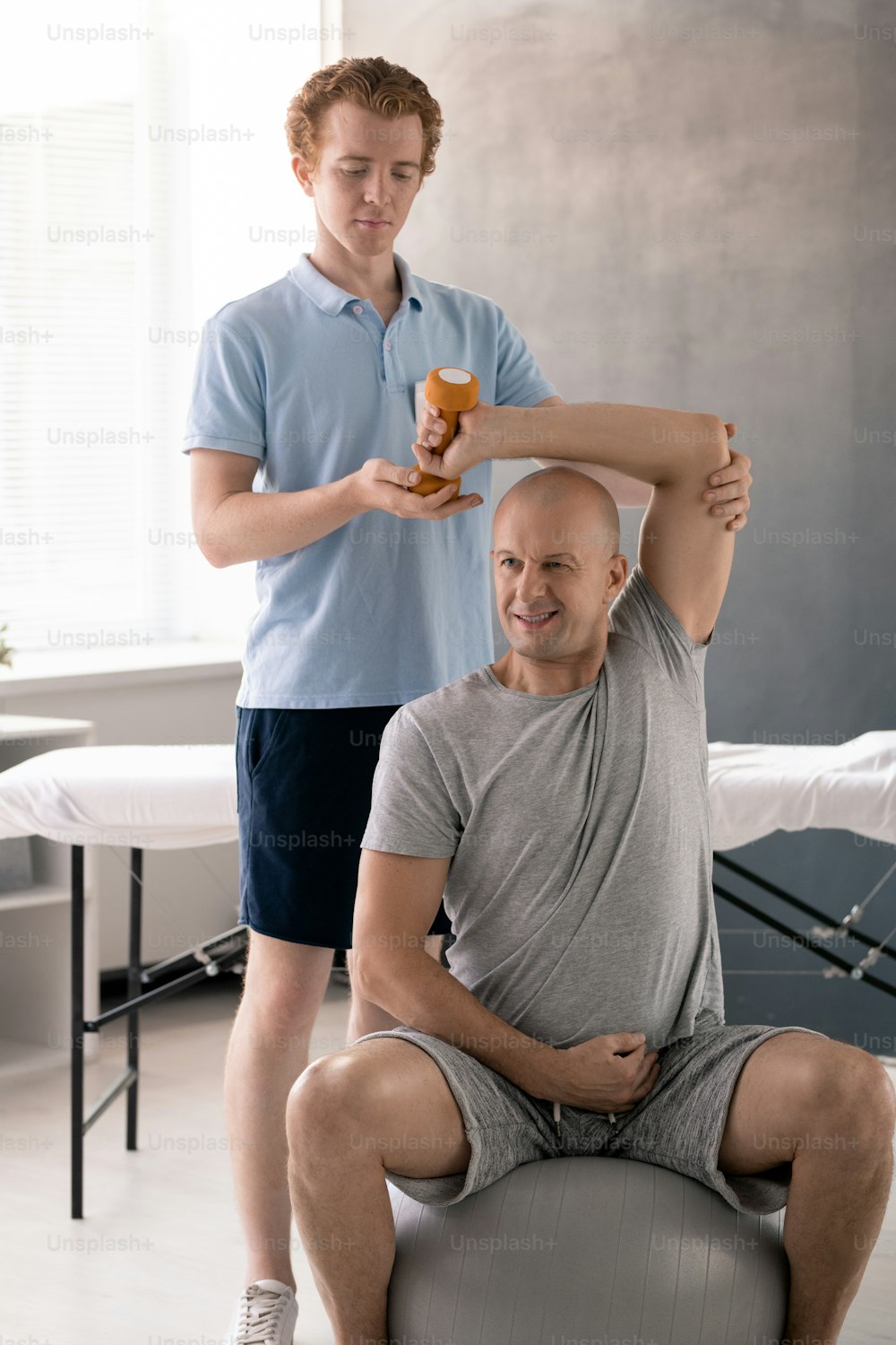 Mature sportsman in activewear sitting on fitball in rehabilitation clinics and doing exercise with dumbbell while clinician helping him