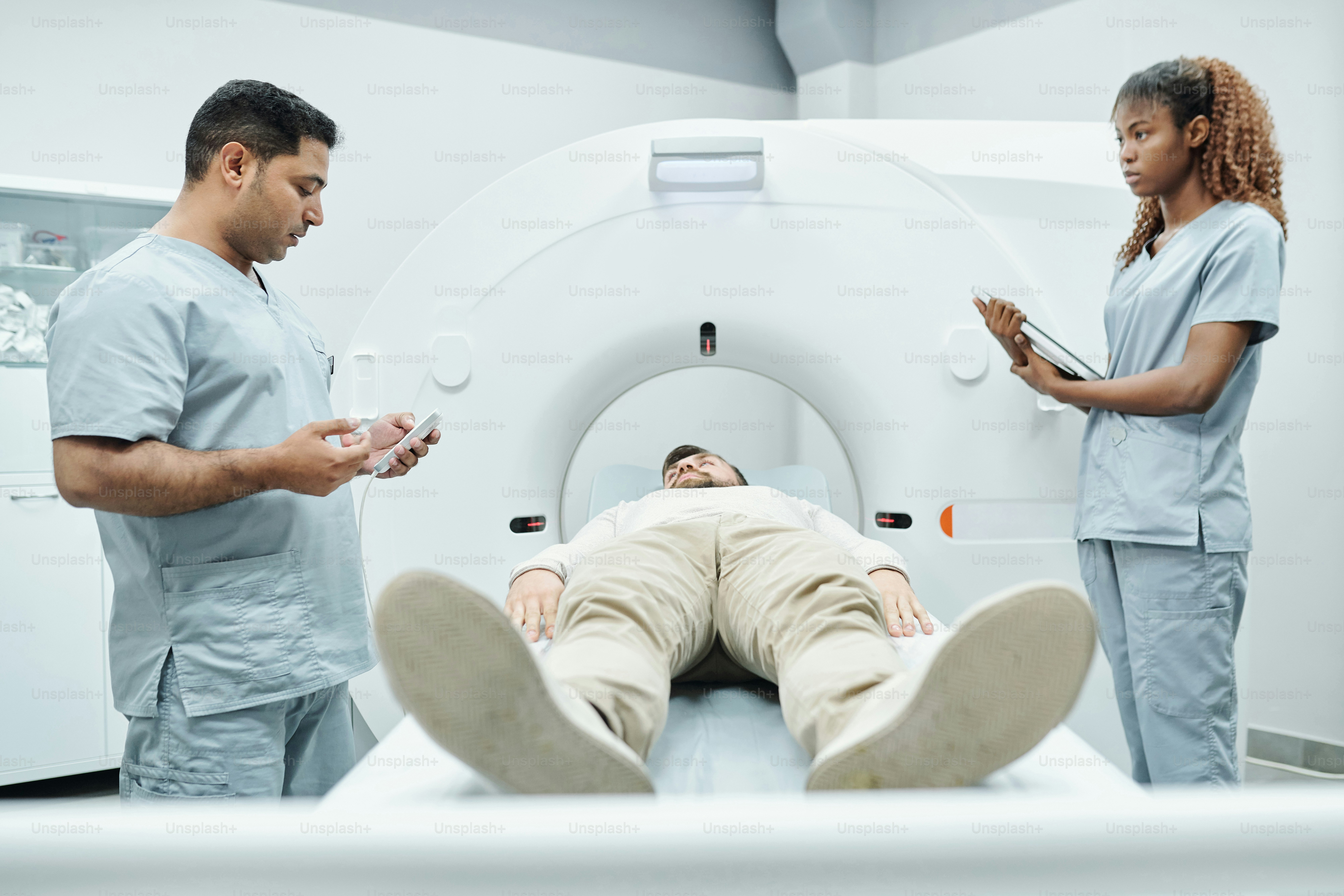 Junge afrikanische Krankenschwester oder Assistentin in Uniform schaut auf einen Arzt, der auf der anderen Seite eines langen Liegeplatzes steht, in dem ein männlicher Patient im medizinischen Zentrum liegt