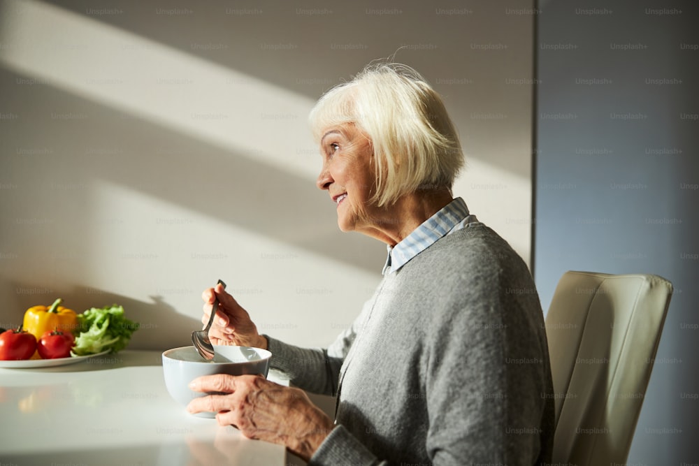 Vista lateral de uma bela mulher loira envelhecida sorridente tomando café da manhã na mesa da cozinha