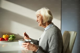 Vista lateral de una hermosa mujer rubia sonriente y envejecida desayunando en la mesa de la cocina