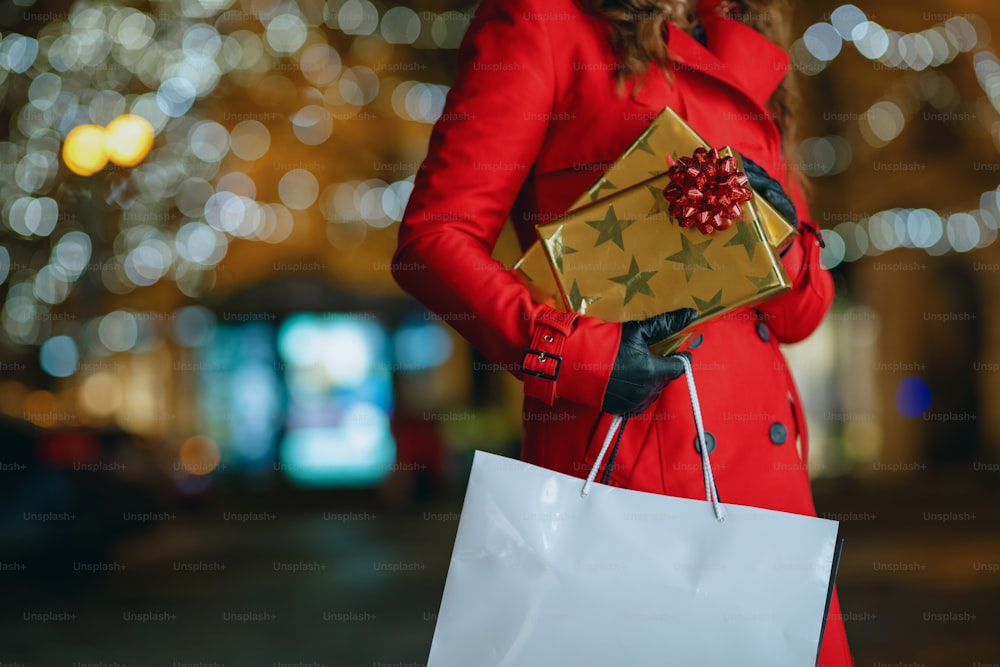 Winterspaß. Nahaufnahme einer Reisenden mittleren Alters in rotem Mantel und schwarzer Baskenmütze mit Einkaufstüten und Geschenken im Freien in der Stadt am Abend.