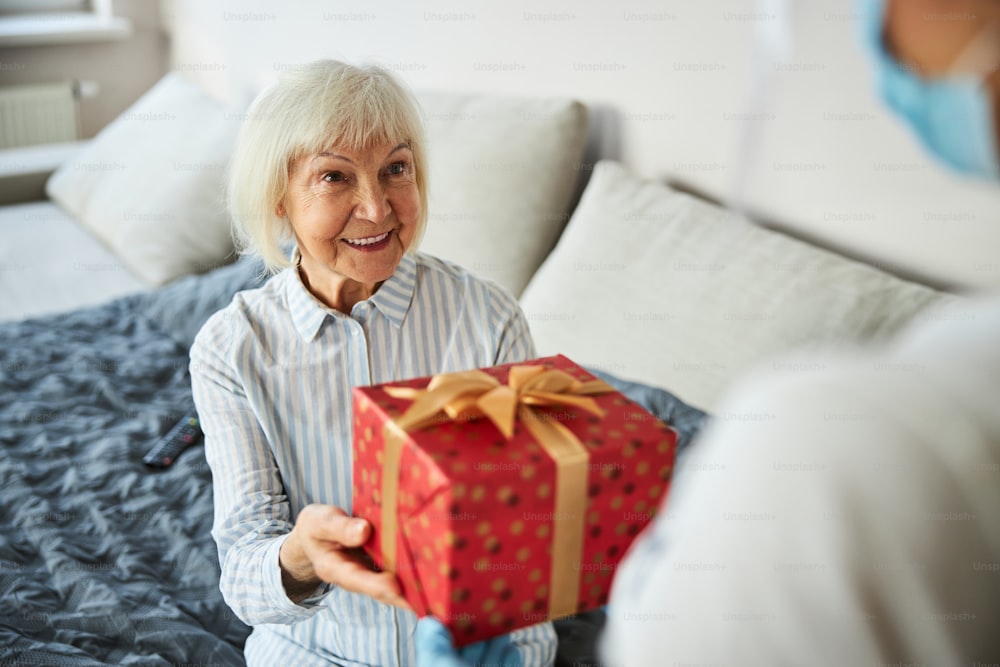 Pleasantly surprised aging person seizing a box inside a wrapping paper from a home care female