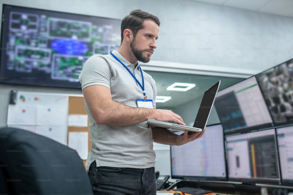 Trabalhando com informação. Homem adulto jovem sério em pé com laptop em escritório especialmente equipado no local de trabalho perto de monitores
