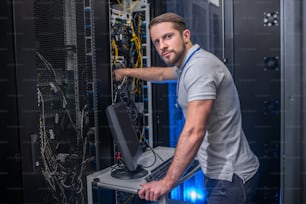 Search, solution. Serious smart young bearded man with badge working in server room checking cable