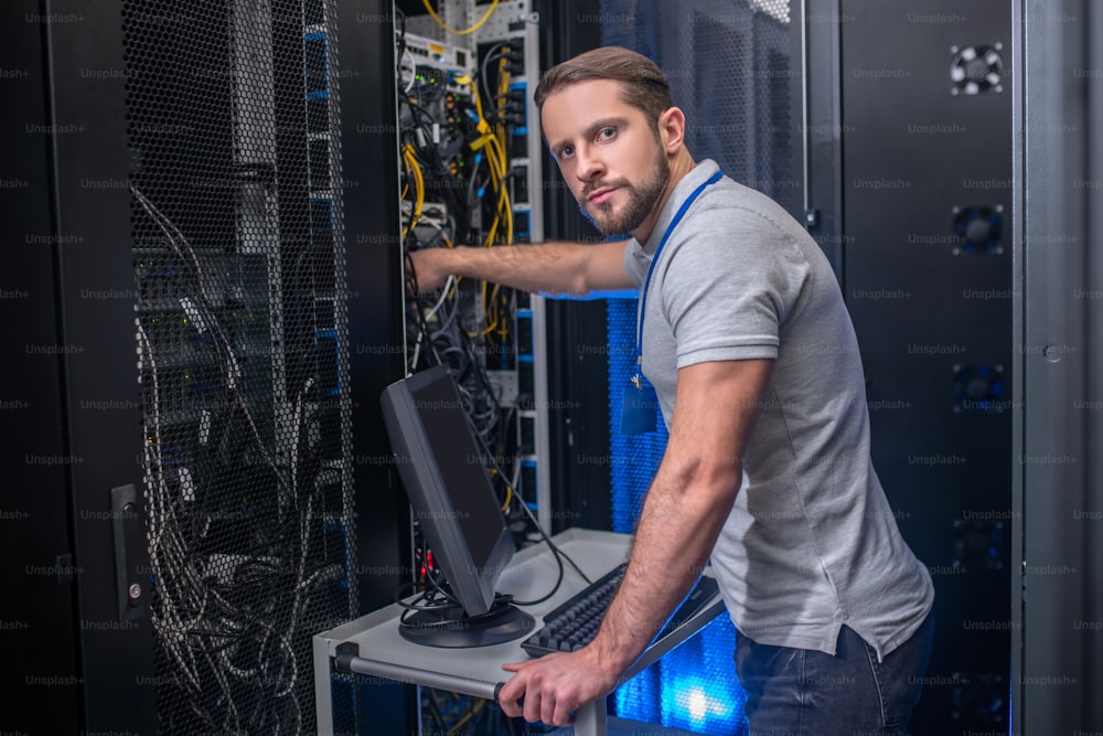 Search, solution. Serious smart young bearded man with badge working in server room checking cable