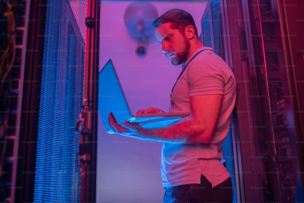 システム管理者。青とピンクの光を持つサービングルームにノートパソコンを持つ若い男性のソフトウェアスペシャリストが立つ