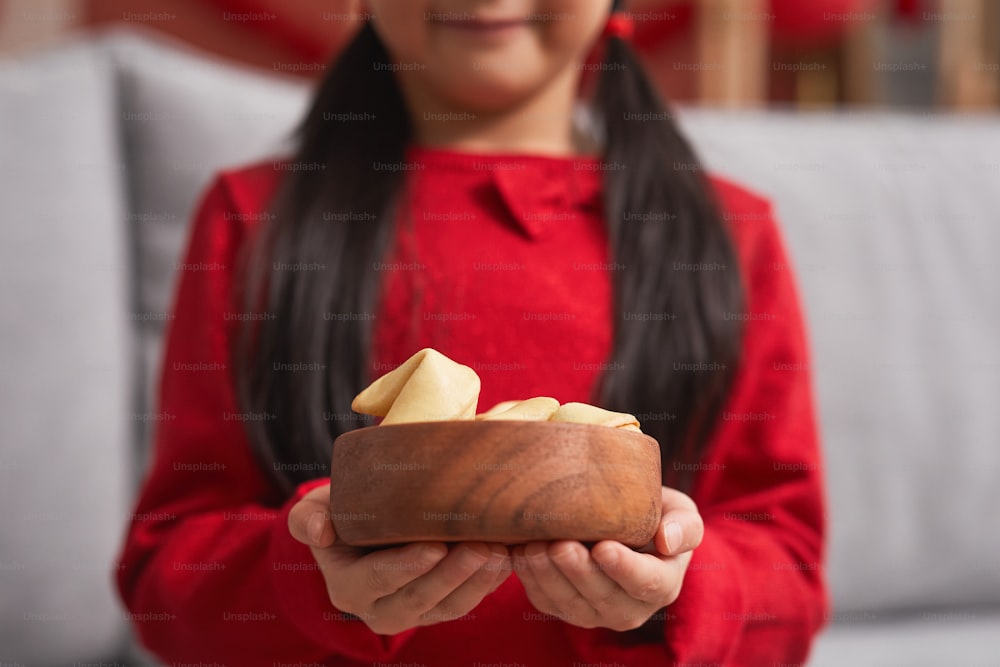 Primer plano de la sección media de una niña asiática irreconocible vestida de rojo con dos colas de caballo sosteniendo un cuenco de madera lleno de galletas de la fortuna