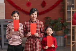 Family portrait of beautiful mature Chinese woman and her daughters standing in living room holding red envelopes, Lunar New Year concept