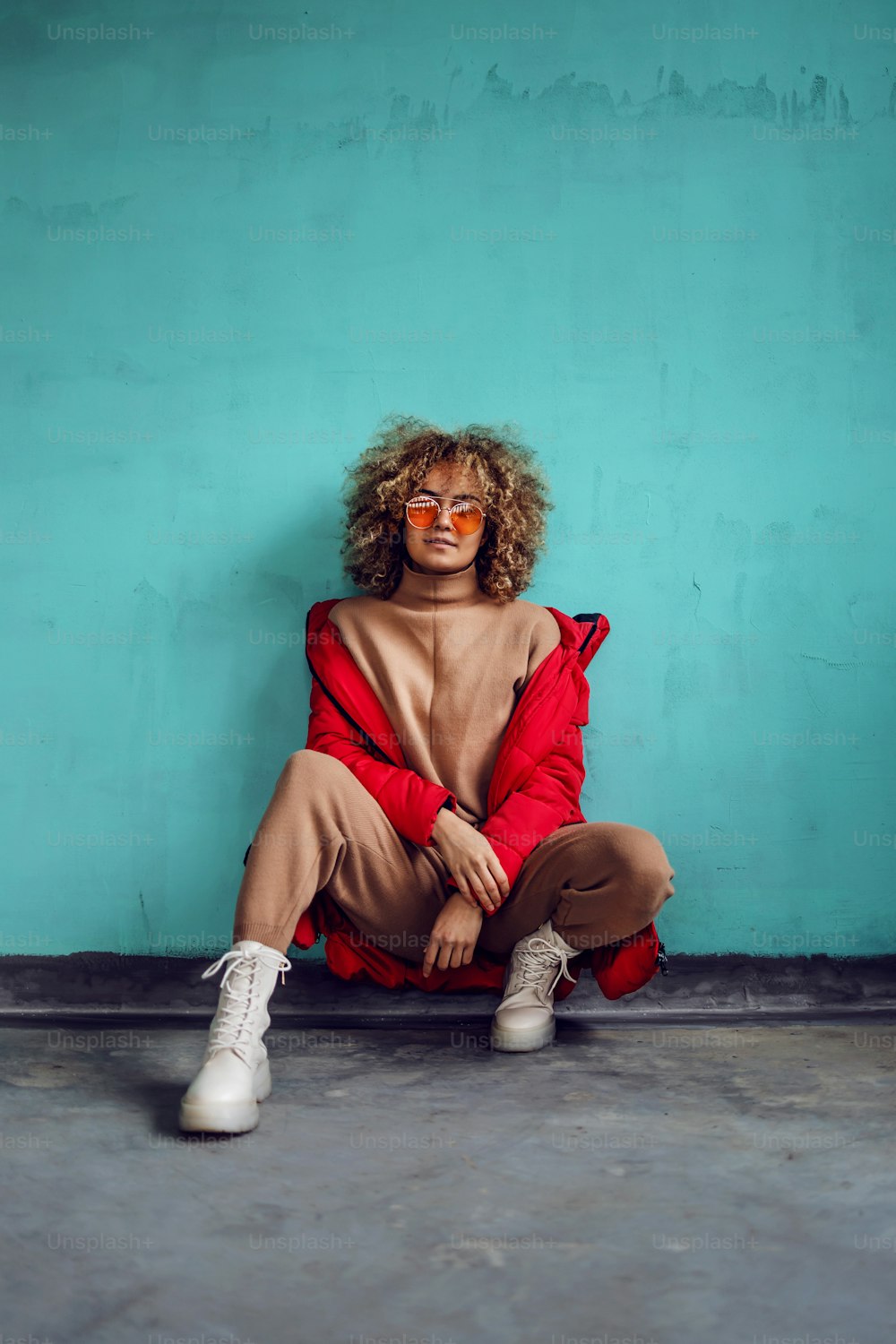 Young fashionable woman with curly hair, in red jacket crouching in front of the wall and looking at camera.