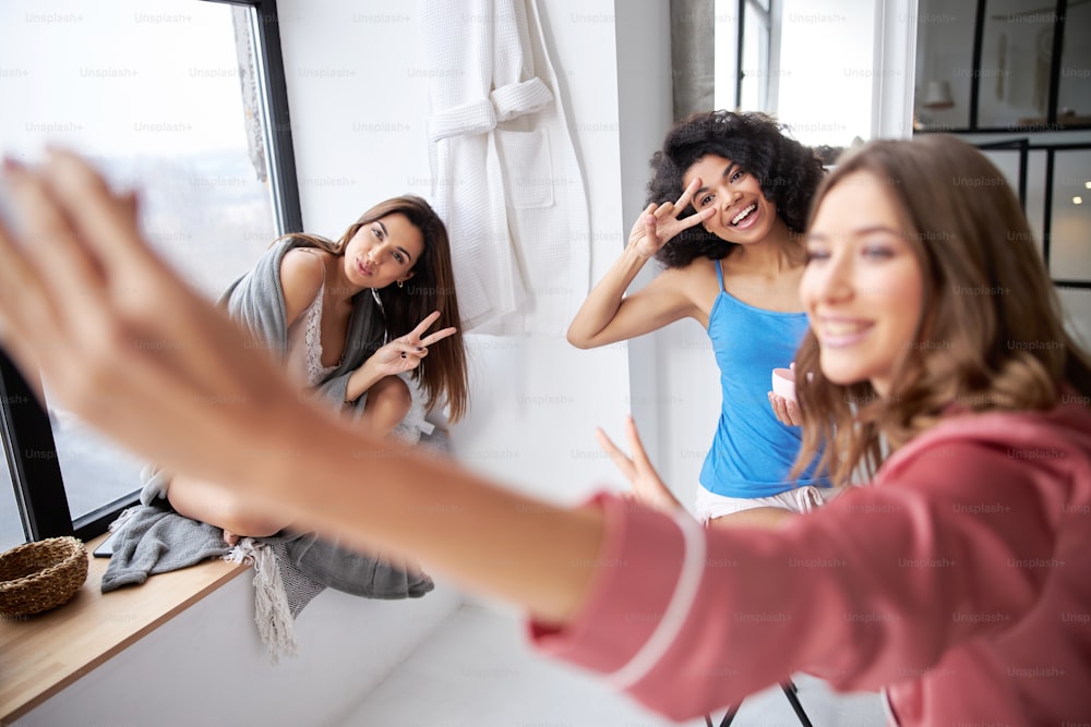Focus on the two cute girls gesturing while making selfie. High angle view of three best friends making funny faces while posing at the camera. Photographing girl is blurred