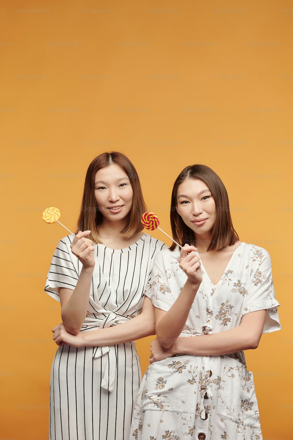 Happy young female twins of Asian ethnicity with lollipops looking at you with smiles while standing in front of camera against yellow background