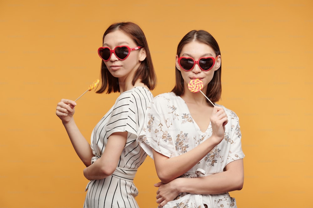Young stylish female twins in smart dresses and sunglasses having lollipops while standing in front of camera against yellow background