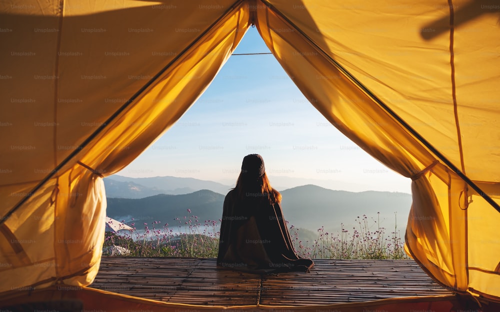 Imagem da vista traseira de uma mulher sentada na varanda de madeira enquanto observa uma bela vista das montanhas e da natureza fora da tenda