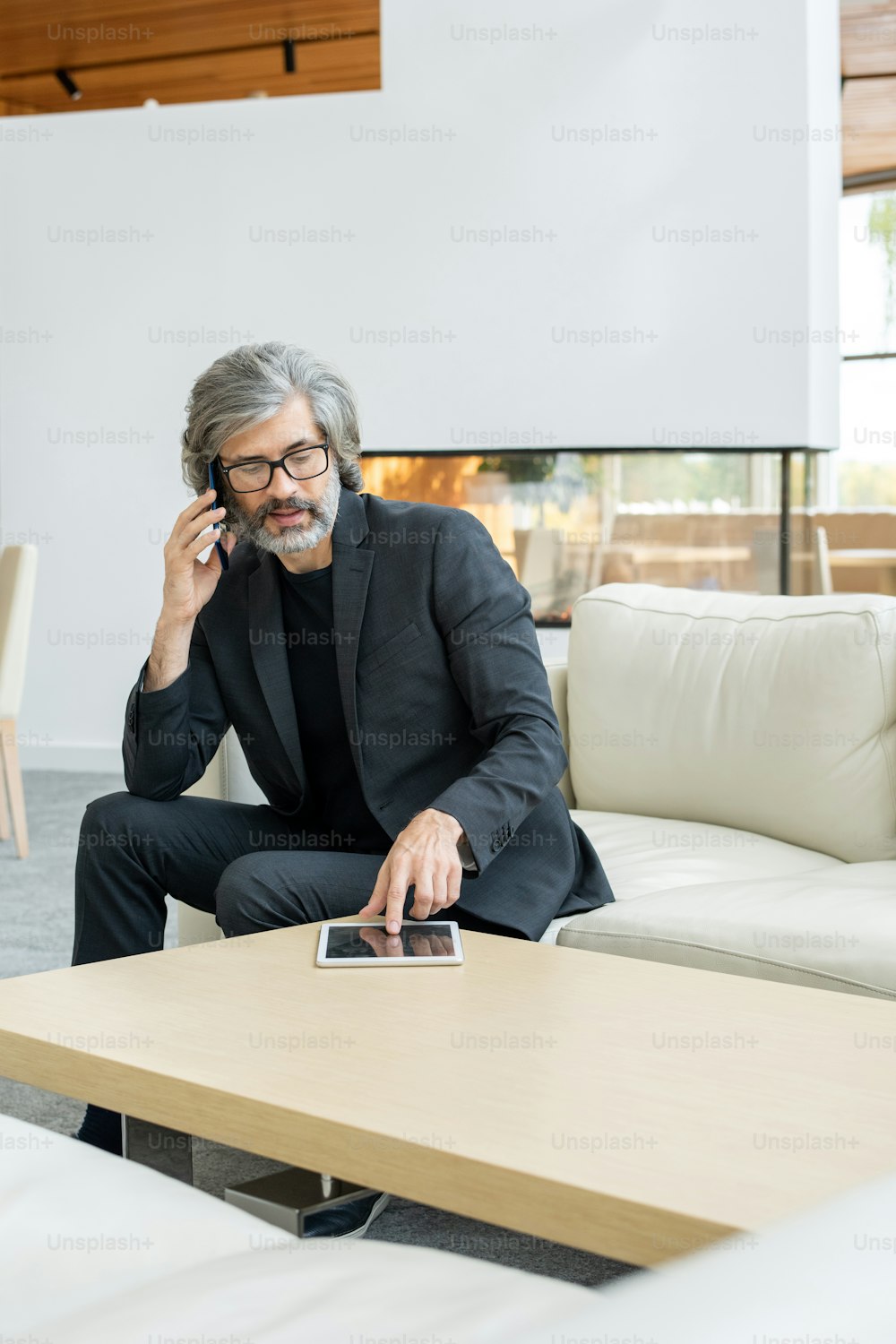 Elegant mature businessman sitting on white leather couch in luxurious restaurant and scrolling in tablet while talking on smartphone