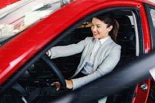 Lächelnde Frau, die im Auto im Autosalon sitzt und ein Auto ausprobiert, das sie kaufen möchte.