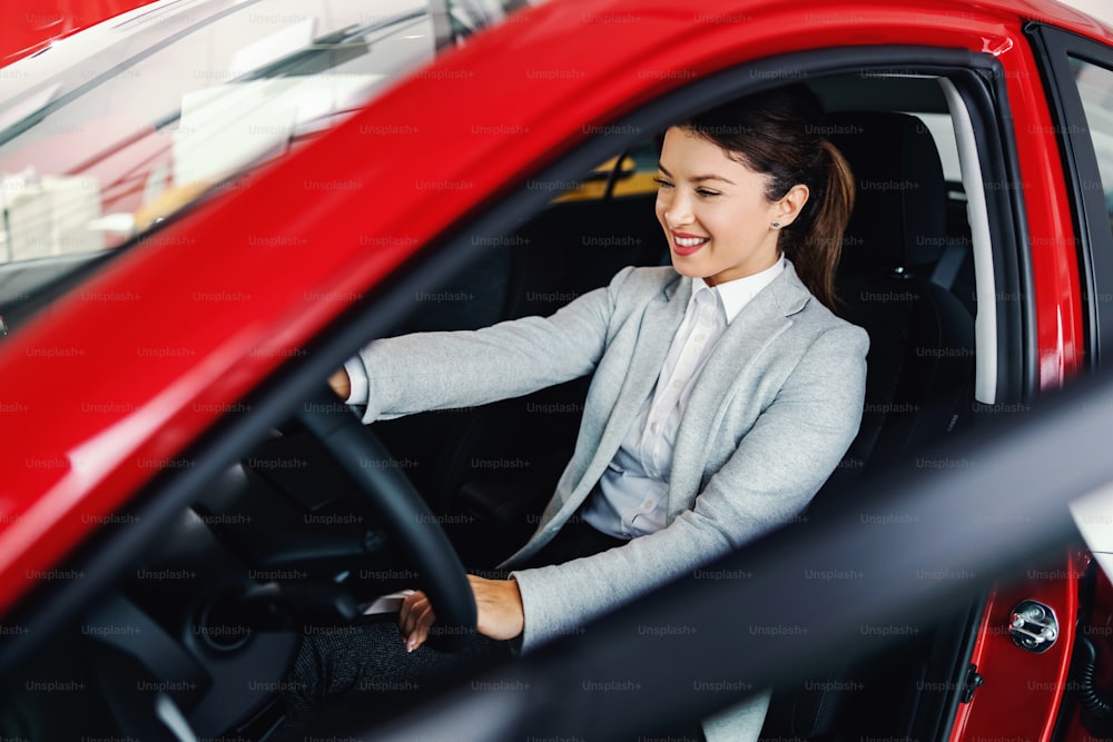 Mujer sonriente sentada en el auto en el salón de autos y probando el auto que quiere comprar.