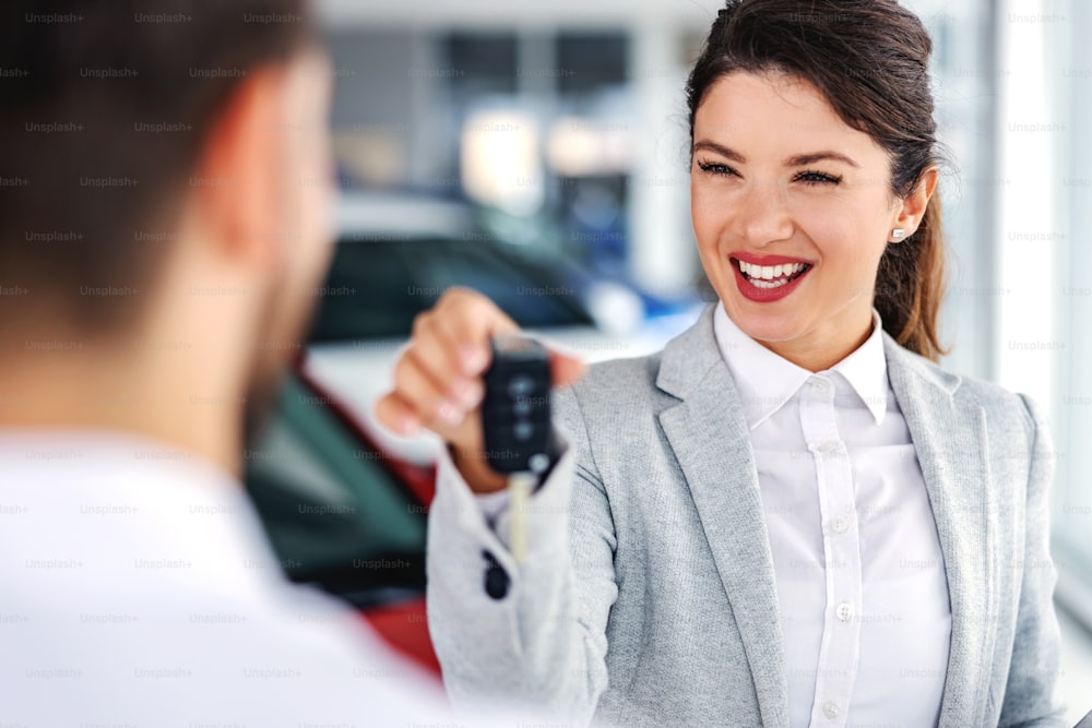 Smiling, friendly car seller standing in car salon with a customer and handing him car keys of best selling car.