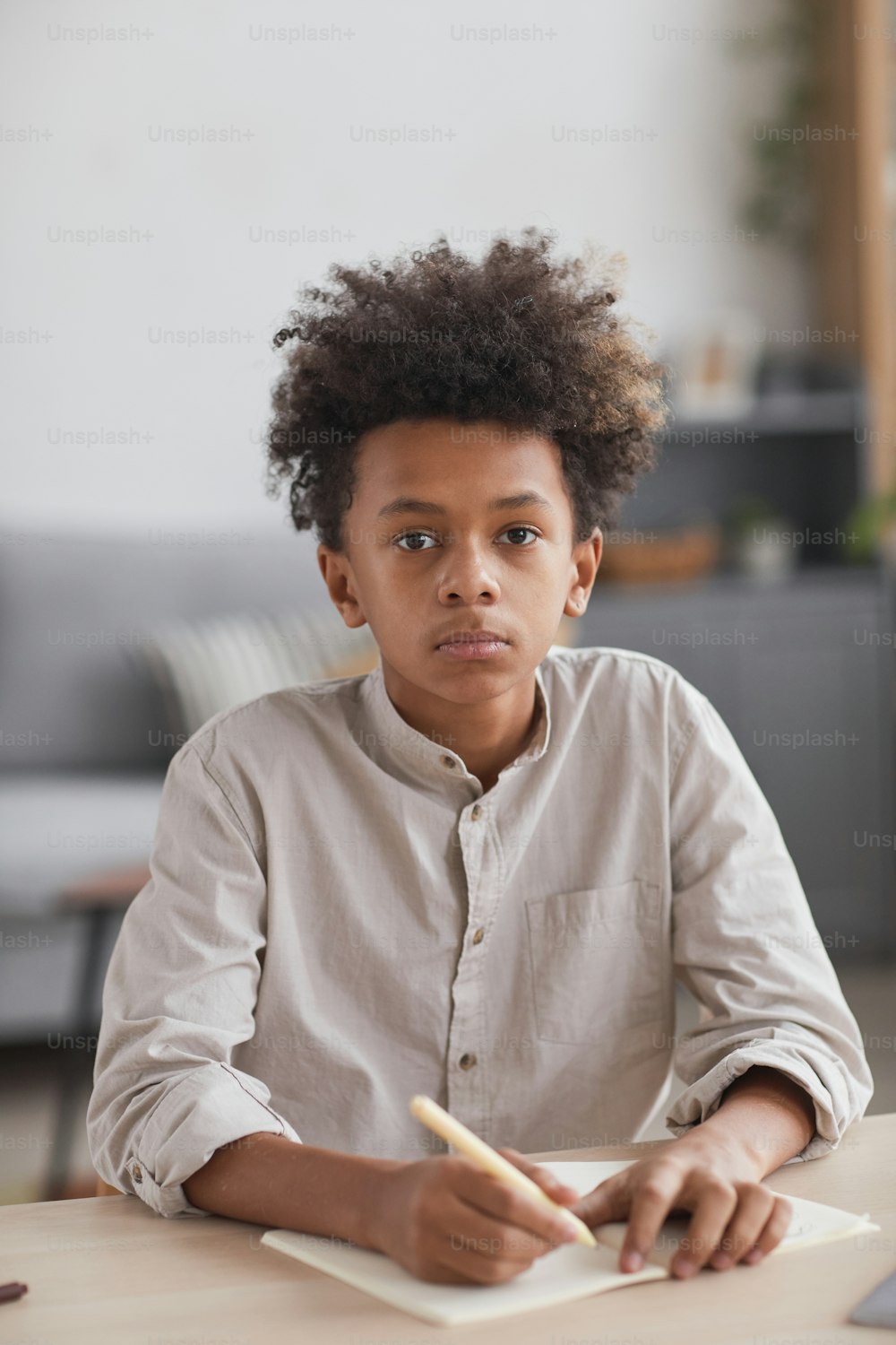 Retrato vertical da vista frontal do menino afro-americano adolescente fazendo a lição de casa na mesa e olhando para a câmera no interior moderno da casa