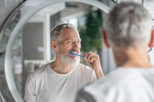 Rotina matinal. Um homem de cabelos grisalhos escovando os dentes perto do espelho