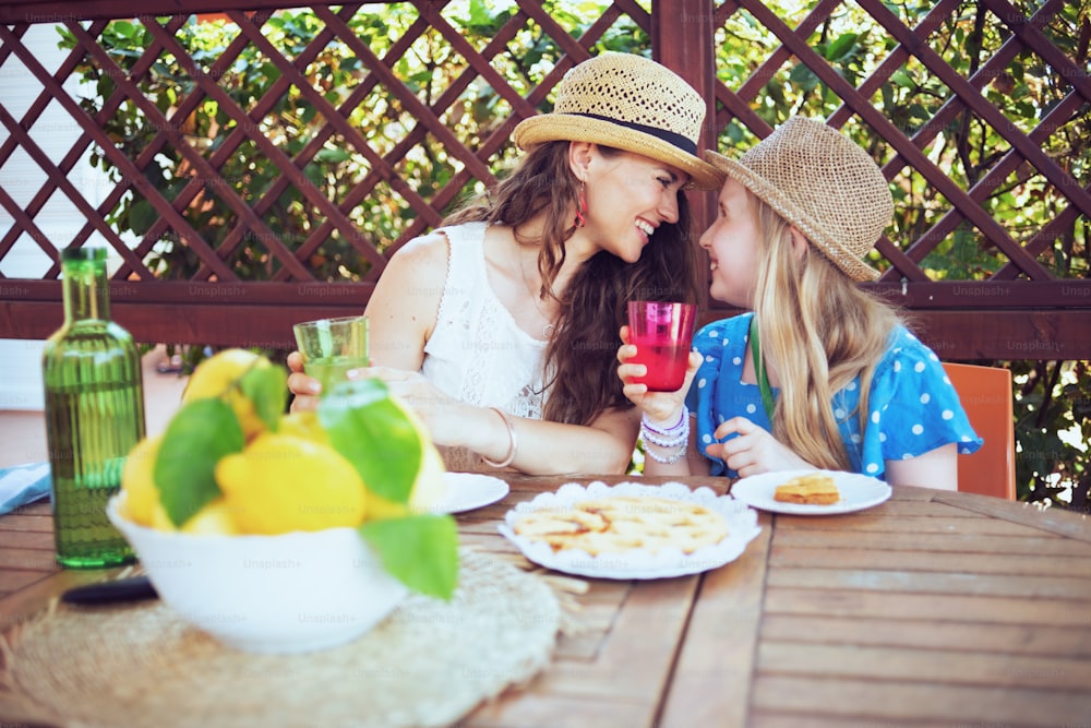 Felice mamma e figlia alla moda con crostata, bottiglia d'acqua verde e piatto di limoni di fattoria locale seduti al tavolo a pranzo nel patio.