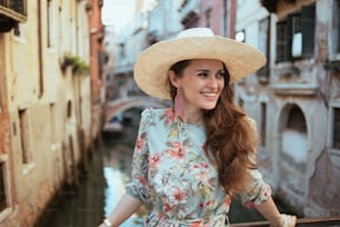 모자를 쓴 꽃무늬 드레스를 입은 행복한 트렌디한 여성이 이탈리아 베니스의 명소를 탐험한다.