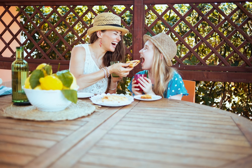緑のレモネードのボトルと地元の農場のレモンのプレートがテラスで朝食をとるテーブルに座っている幸せなスタイリッシュな家族。