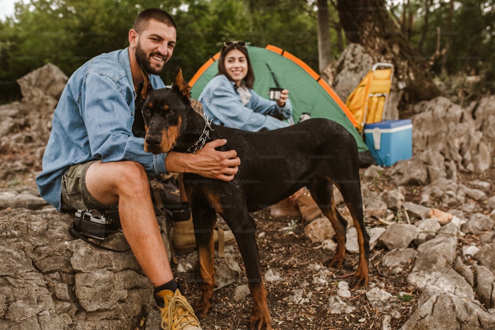 Man and woman and their doberman sitting in chairs outside the tent having coffee and talking. Couple camping.