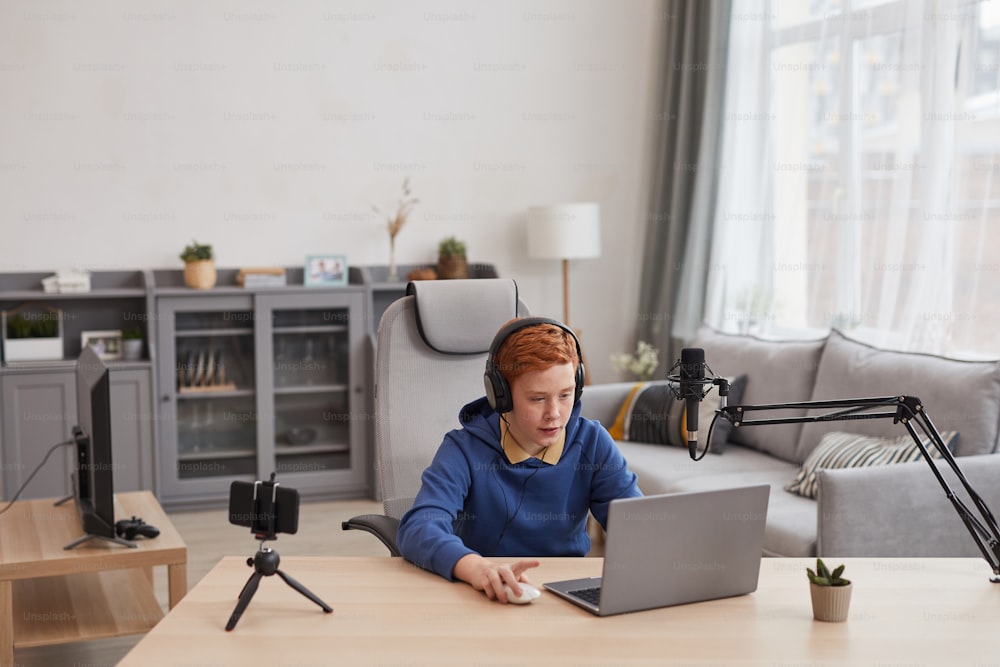 Wide angle portrait of red haired teenage boy playing video games with microphone and camera set up for online streaming, copy space