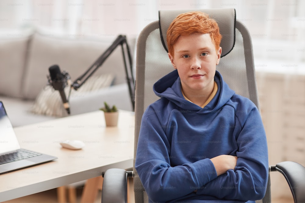 Retrato de un adolescente pelirrojo mirando a la cámara mientras está sentado en una silla contra la computadora en el fondo, espacio de copia