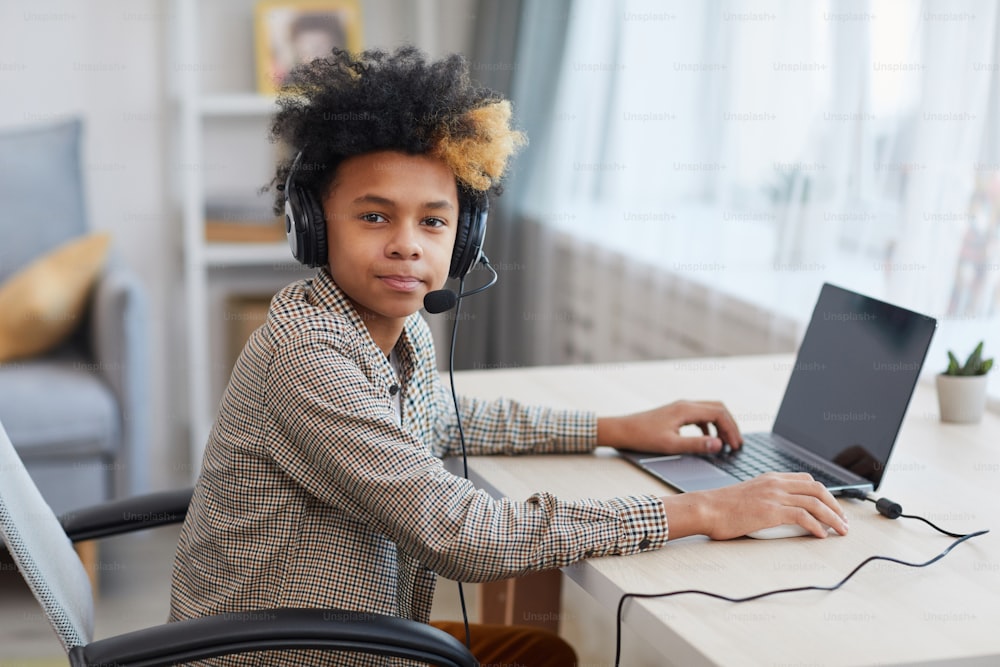 Ritratto di ragazzo afro-americano adolescente che indossa l'auricolare e guarda la fotocamera mentre utilizza il laptop a casa, concetto di giovane giocatore o blogger, spazio di copia