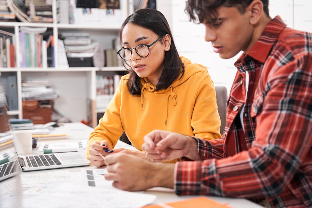 Important negotiations. Portrait view of male and female designers wearing casual clothes are sitting at the table at the office and talking together while looking at the sketches