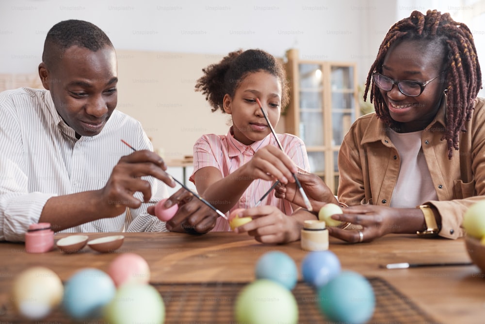 Portrait of happy African-American family painting Easter eggs together while sitting at wooden table in cozy home interior, DIY Easter decorations