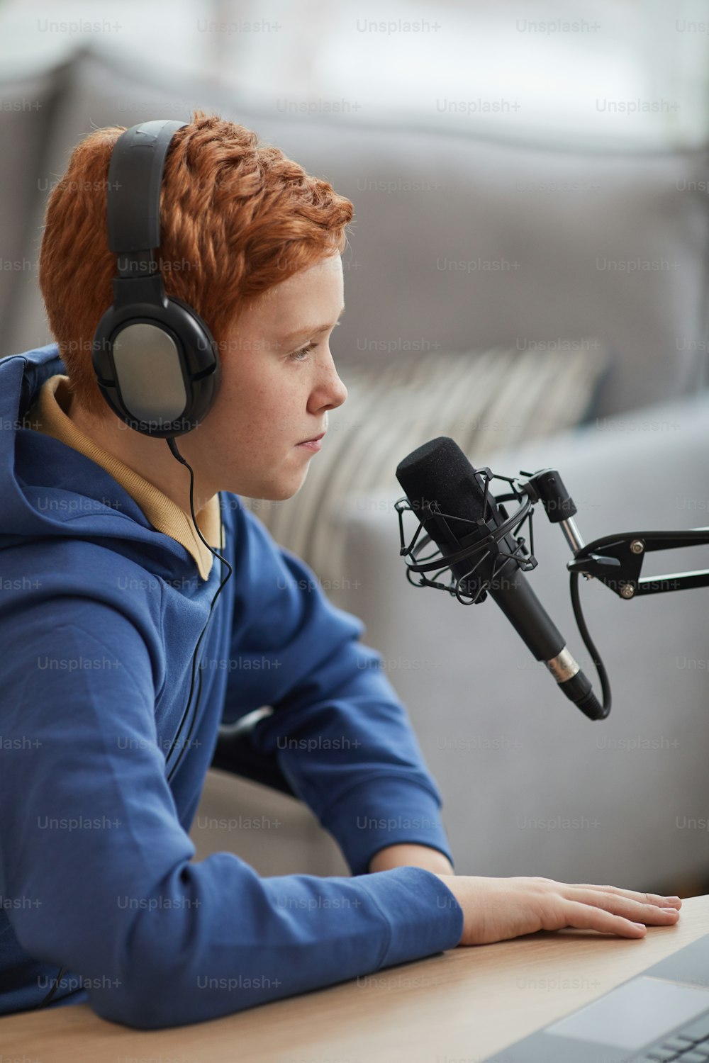 Vertical side view portrait of red haired teenage boy speaking to microphone and wearing headphones while recording podcast or online stream