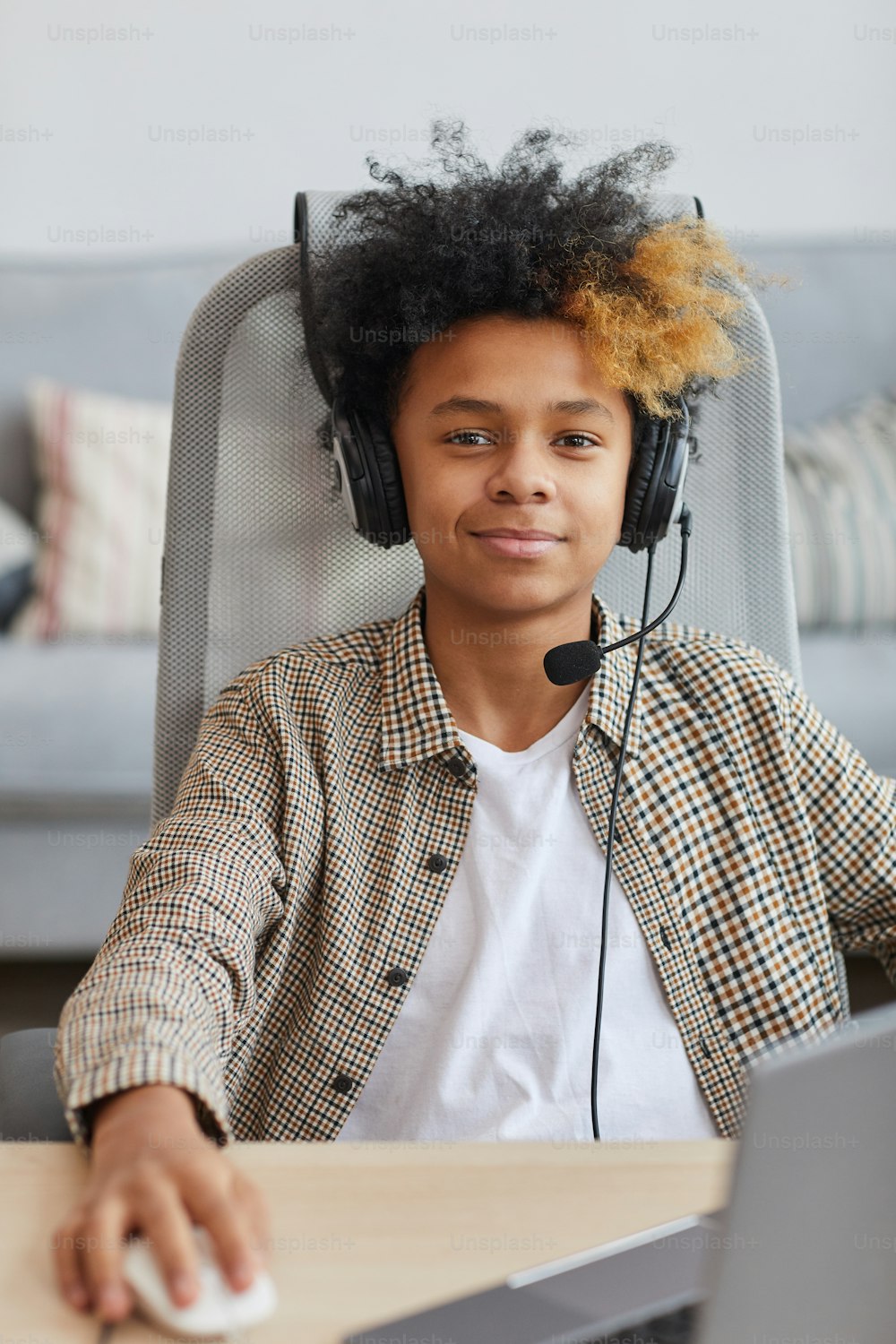 Vertikales Porträt eines lächelnden afroamerikanischen Jungen, der ein Headset trägt, während er zu Hause einen Laptop benutzt und in die Kamera schaut