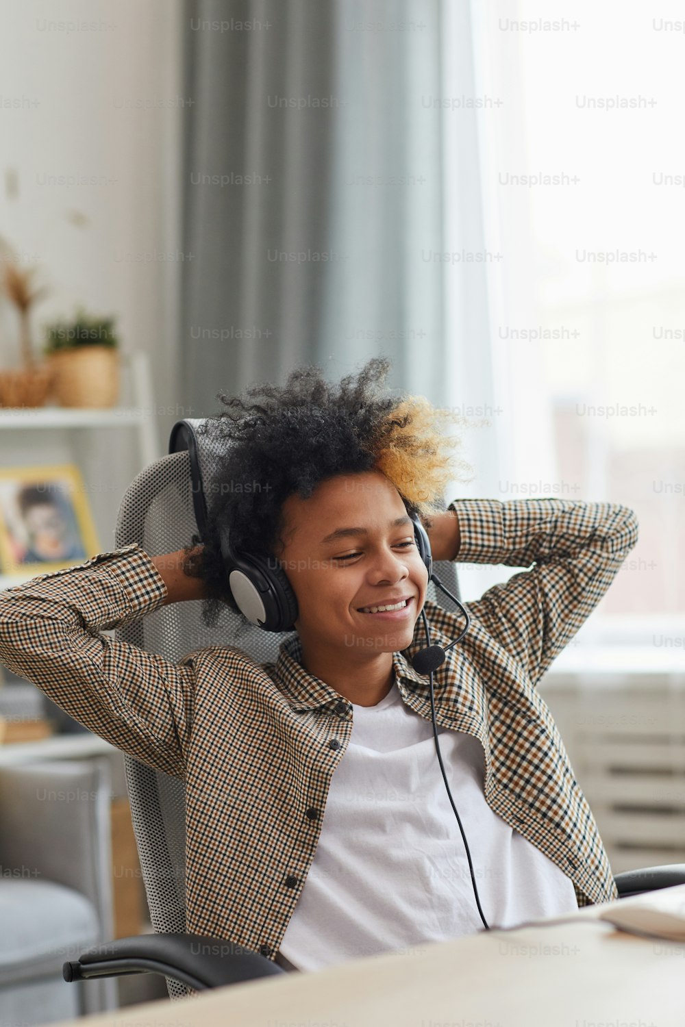 Ritratto verticale di sorridente ragazzo afro-americano che indossa l'auricolare e si rilassa sulla sedia mentre utilizza il laptop a casa, concetto di giocatore o blogger