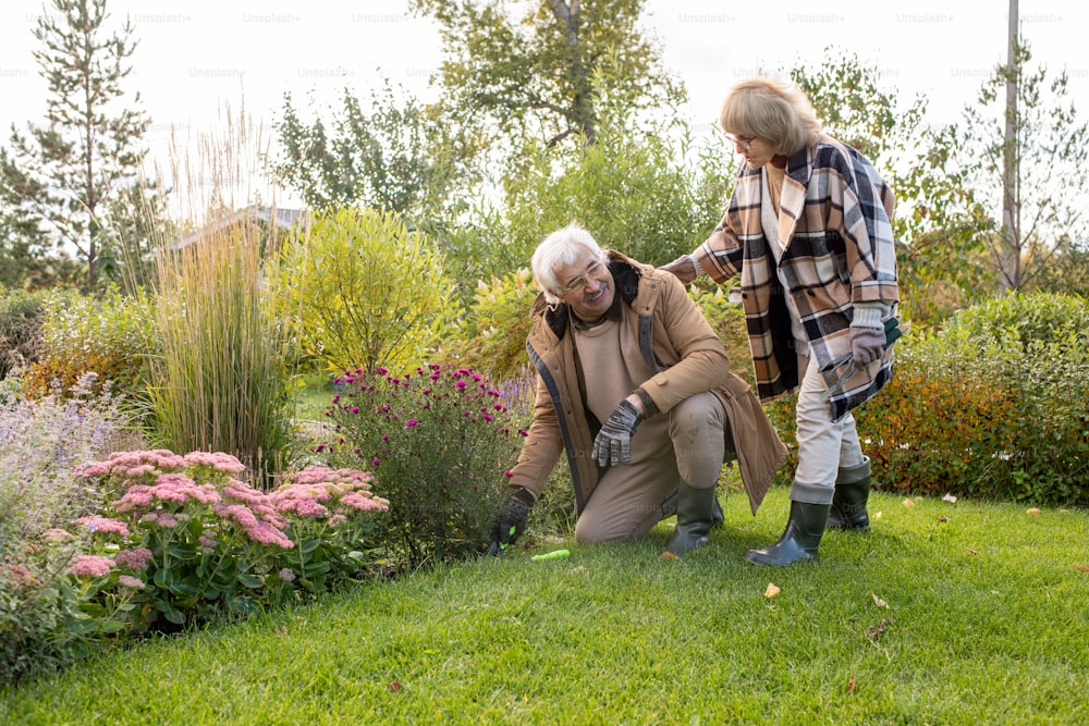 Retired blond female in eyeglasses and warm stylish checkered coat helping her husband standing on knees on green lawn by flowerbed