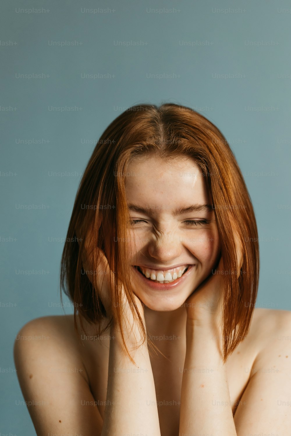Laughing vivacious young redhead woman with a beaming smile holding her hands near her face while posing over grey background. Happy woman with naked shoulders laughing with downcast