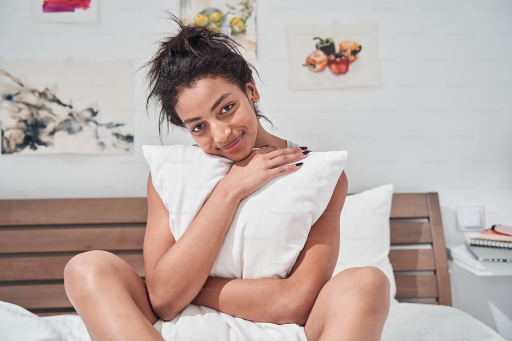 Portrait view of the multiracial woman just wake up in the morning and relaxing on the bed. She open the window to receive the light of the morning sun. Girl hugging a pillow on her bed in the bedroom