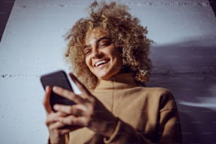Une joyeuse fille de rap métisse debout contre le pilier et utilisant un téléphone intelligent pour envoyer des SMS à un ami ou accrocher sur les médias sociaux.