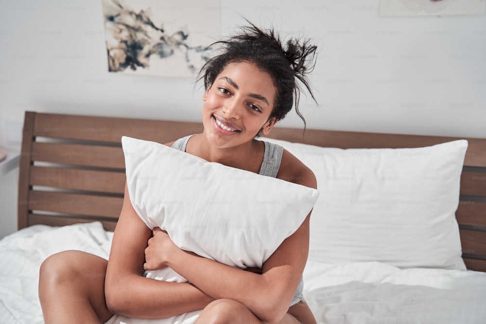 Young beautiful girl smiling while sitting in bed and hugging pillow. Multiracial girl smiling to the camera while relaxing at the cozy bedroom. Morning concept. Stock photo