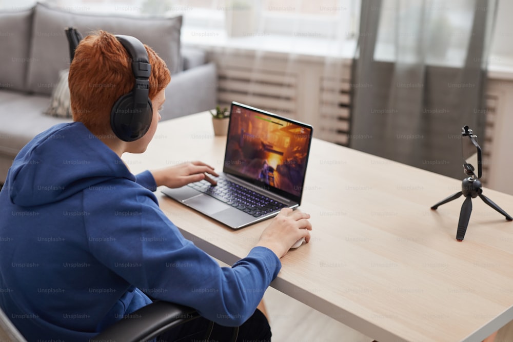 Vue arrière portrait d’un adolescent aux cheveux roux jouant à des jeux vidéo avec des écouteurs et une caméra configurés pour le streaming en ligne, espace de copie