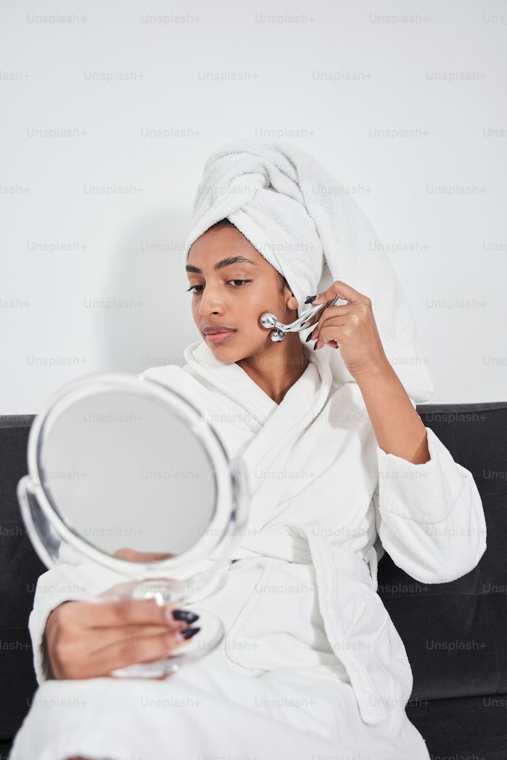 Portrait view of brunette attractive female wearing bathrobe is holding mirror and looking into it while doing face massage indoors. Concept of beauty, self-care, cosmetics