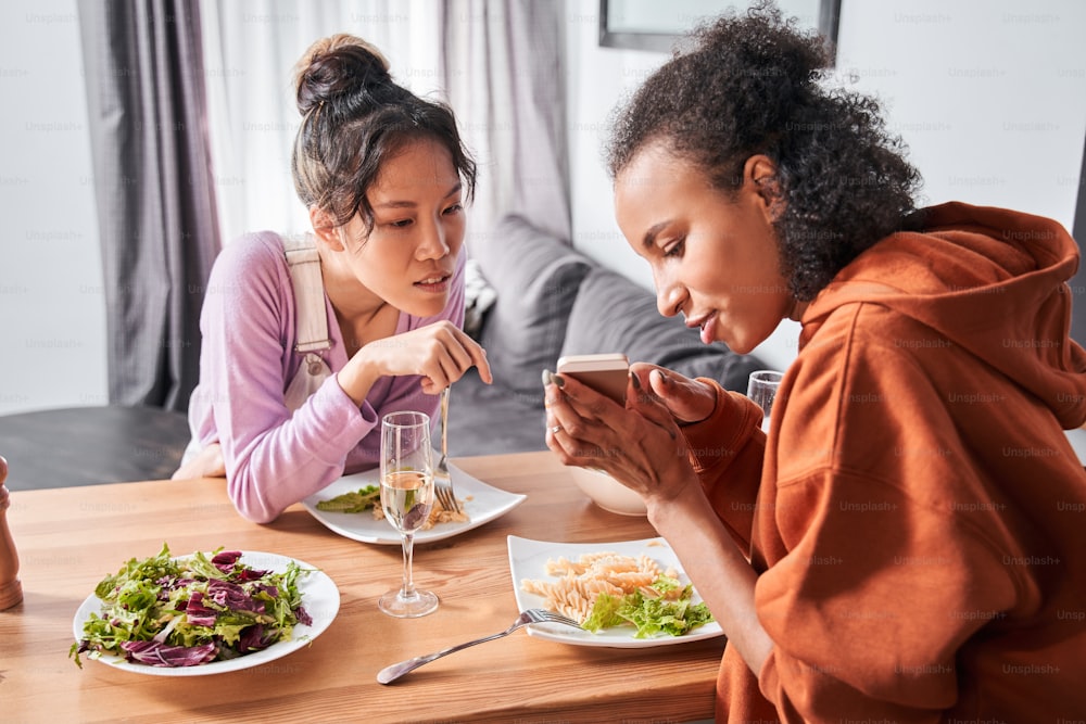 Portrait view of the couple of multiracial friends sitting at the bar counter and having dinner at the kitchen. Curly girl showing something at the smartphone to her bestie. Stock photo