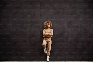 Chica de hip hop en chándal parada contra la pared con los brazos cruzados.