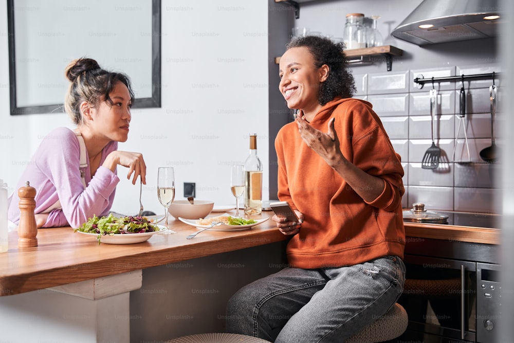 Pasta and conversations. Cute multiracial woman laughing while telling a funny story to her bestie at the glass of wine. Two happy girls sitting at the kitchen and eating tasty pasta