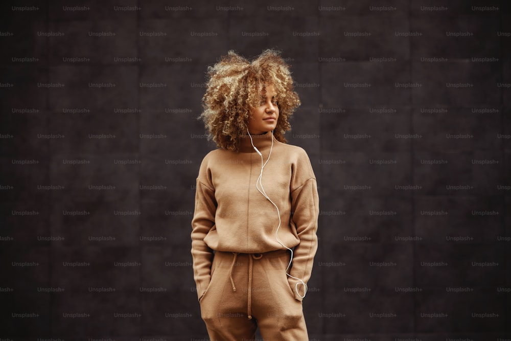 Hip hop girl in tracksuit standing with hands in her pockets, listening music and looking away.