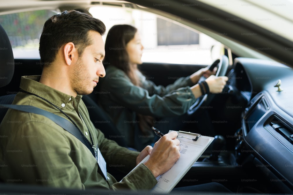 Serious male instructor writing and taking notes during a driving class with an adolescent girl. Man in his 30s grading the driving skills of a teenage girl