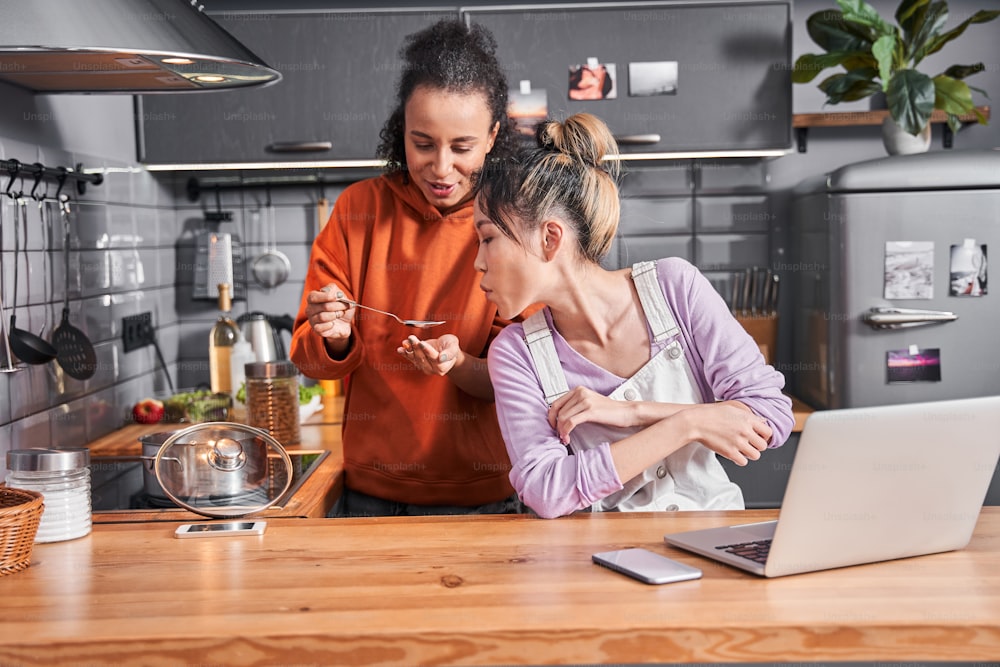 Try this one. Portrait view of the multiracial woman gives a taste her dish to her best friend while cooking together at the kitchen. Coolinary concept